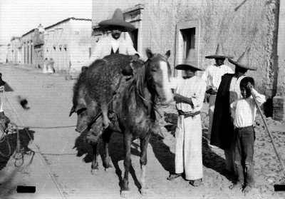 Hombre a caballo transporta cerdo muerto por calle de un poblado, retrato