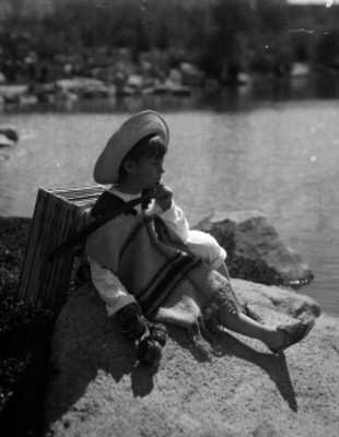 Niño con un huacal en la espalda y tarros en una mano, sentado sobre una piedra a la orilla de un lago, retrato