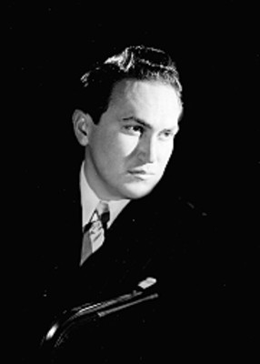 Nicolás Urcelay, cantante de opera, sentado de perfil en una silla, viste de traje a rayas y corbata, retrato