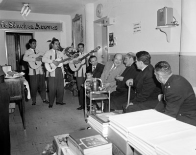 Sacerdote sentado junto a otros hombres escuchan cantar a un trío musical en la sala de espera de un consultorio